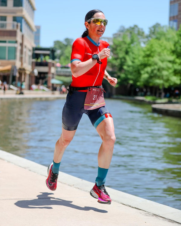 Athlete Amber Ferriera running along a waterfront path with buildings and trees in the background in a triathlon kit and a race bib on her lower stomach.